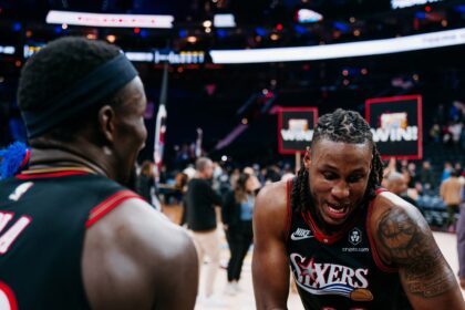 Justin Edwards #33 of the Philadelphia 76ers, showing a large tattoo on his left shoulder, grips a basketball and smiles while celebrating with a teammate whose back is to the camera.