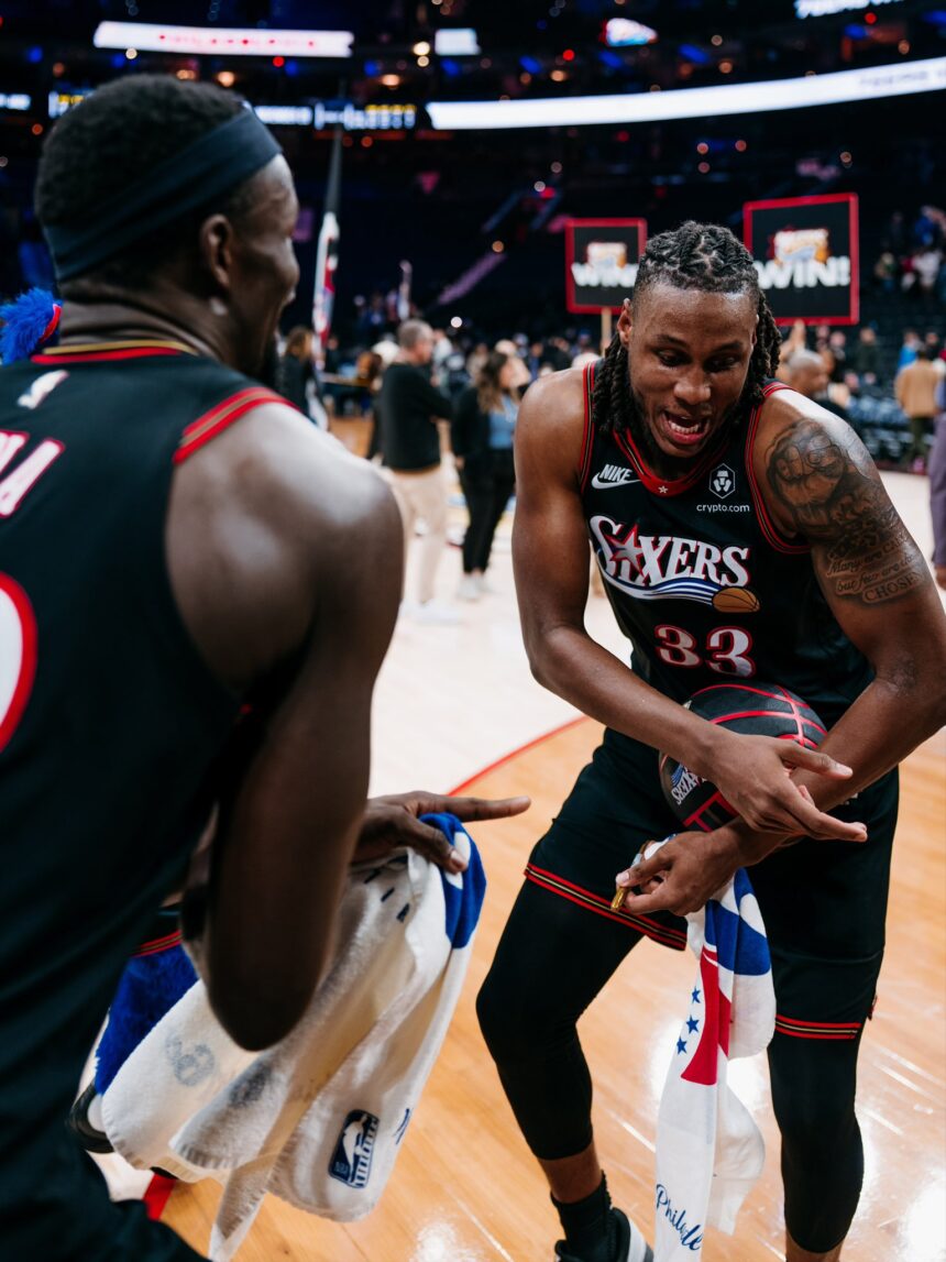 Justin Edwards #33 of the Philadelphia 76ers, showing a large tattoo on his left shoulder, grips a basketball and smiles while celebrating with a teammate whose back is to the camera.