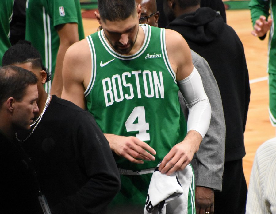 A medium shot of Boston Celtics player Nikola Vucevic (wearing jersey number 4) looking down with a somber expression while holding a towel. He is on the court surrounded by staff and teammates during a game