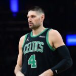 A medium shot of Boston Celtics center Nikola Vučević (wearing jersey number 4) looking off-camera with a serious expression. He is wearing a black Celtics "Statement Edition" jersey with green trim.