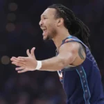 A close-up vertical shot of NBA player Jaylin Williams of the Oklahoma City Thunder. He is wearing the team's "Icon Edition" blue jersey with "OKC" across the chest in white and orange. His mouth is open in a wide shout of celebration or intensity, and his arms are partially visible and spread wide. The background shows the blurred bright lights and dark rafters of a professional basketball arena.
