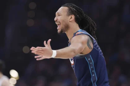 A close-up vertical shot of NBA player Jaylin Williams of the Oklahoma City Thunder. He is wearing the team's "Icon Edition" blue jersey with "OKC" across the chest in white and orange. His mouth is open in a wide shout of celebration or intensity, and his arms are partially visible and spread wide. The background shows the blurred bright lights and dark rafters of a professional basketball arena.