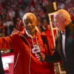 A medium shot of Nolan Richardson, former head coach of the Arkansas Razorbacks, smiling and waving to a crowd during an interview on a basketball court. He is wearing a red jacket with the "Razorbacks" logo and is surrounded by fans in red shirts in the background stands.
