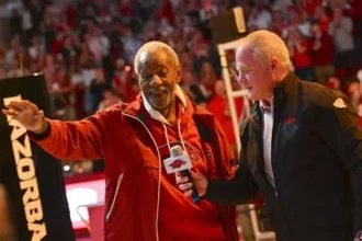 A medium shot of Nolan Richardson, former head coach of the Arkansas Razorbacks, smiling and waving to a crowd during an interview on a basketball court. He is wearing a red jacket with the "Razorbacks" logo and is surrounded by fans in red shirts in the background stands.