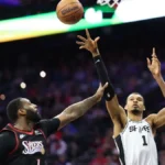 A wide-angle shot of NBA players Victor Wembanyama (San Antonio Spurs) and Andre Drummond (Philadelphia 76ers) during a basketball game. Wembanyama, in a white jersey with the number 1, is jumping with his long right arm fully extended toward the basketball in the air. Drummond, in a black Sixers jersey, is positioned below him, reaching up to contest the play. The background shows a blurred, crowded arena with stadium lights.