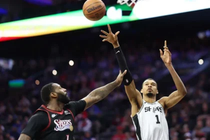 A wide-angle shot of NBA players Victor Wembanyama (San Antonio Spurs) and Andre Drummond (Philadelphia 76ers) during a basketball game. Wembanyama, in a white jersey with the number 1, is jumping with his long right arm fully extended toward the basketball in the air. Drummond, in a black Sixers jersey, is positioned below him, reaching up to contest the play. The background shows a blurred, crowded arena with stadium lights.