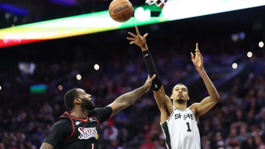A wide-angle shot of NBA players Victor Wembanyama (San Antonio Spurs) and Andre Drummond (Philadelphia 76ers) during a basketball game. Wembanyama, in a white jersey with the number 1, is jumping with his long right arm fully extended toward the basketball in the air. Drummond, in a black Sixers jersey, is positioned below him, reaching up to contest the play. The background shows a blurred, crowded arena with stadium lights.