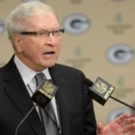 Bob Harlan with white hair and glasses, wearing a black suit and a patterned tie, speaks at a podium. He is positioned in front of a backdrop featuring the Green Bay Packers logo. Two microphones with the NFL Network logo are in front of him.