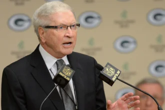 Bob Harlan with white hair and glasses, wearing a black suit and a patterned tie, speaks at a podium. He is positioned in front of a backdrop featuring the Green Bay Packers logo. Two microphones with the NFL Network logo are in front of him.