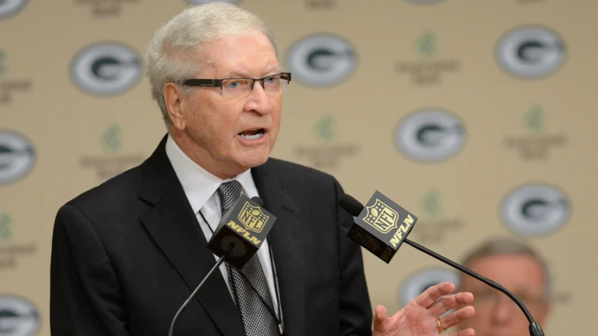Bob Harlan with white hair and glasses, wearing a black suit and a patterned tie, speaks at a podium. He is positioned in front of a backdrop featuring the Green Bay Packers logo. Two microphones with the NFL Network logo are in front of him.