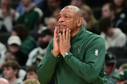 Head coach Doc Rivers, wearing a green Milwaukee Bucks pullover, watches from the sideline with his hands on his face during an NBA game, surrounded by blurred spectators