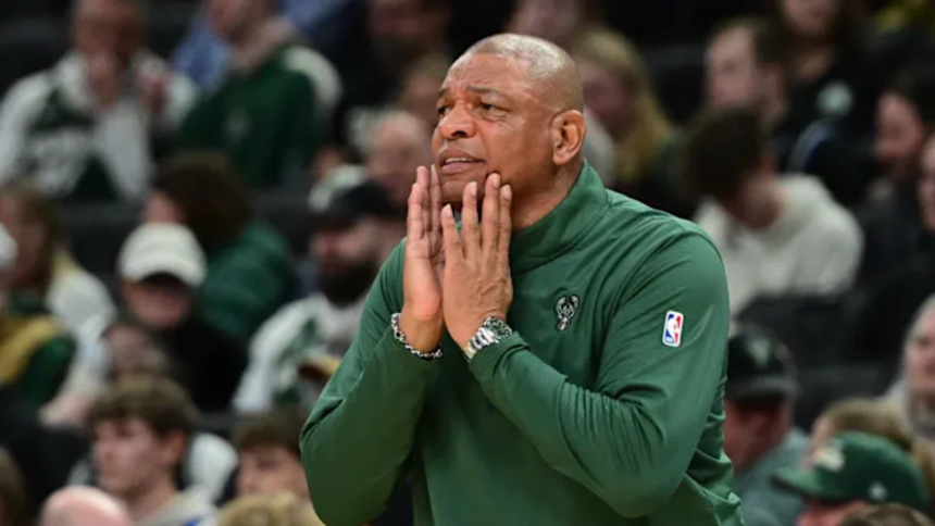Head coach Doc Rivers, wearing a green Milwaukee Bucks pullover, watches from the sideline with his hands on his face during an NBA game, surrounded by blurred spectators
